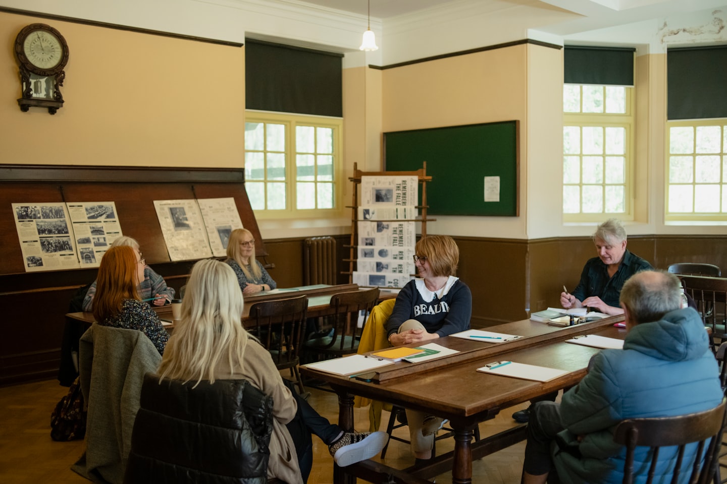 Dospelí diskutujú pri drevených stoloch v staromódnej učebni.